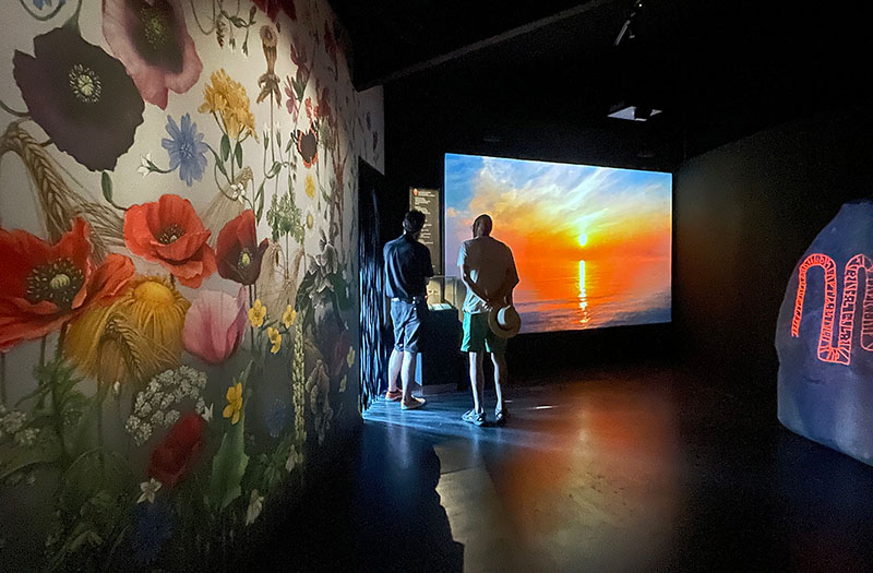 Utställningens sista del med utgång, en stor sten med projicerad runtext, en väggtapet med sensommarblommor och en filmduk som visar en solnedgång.