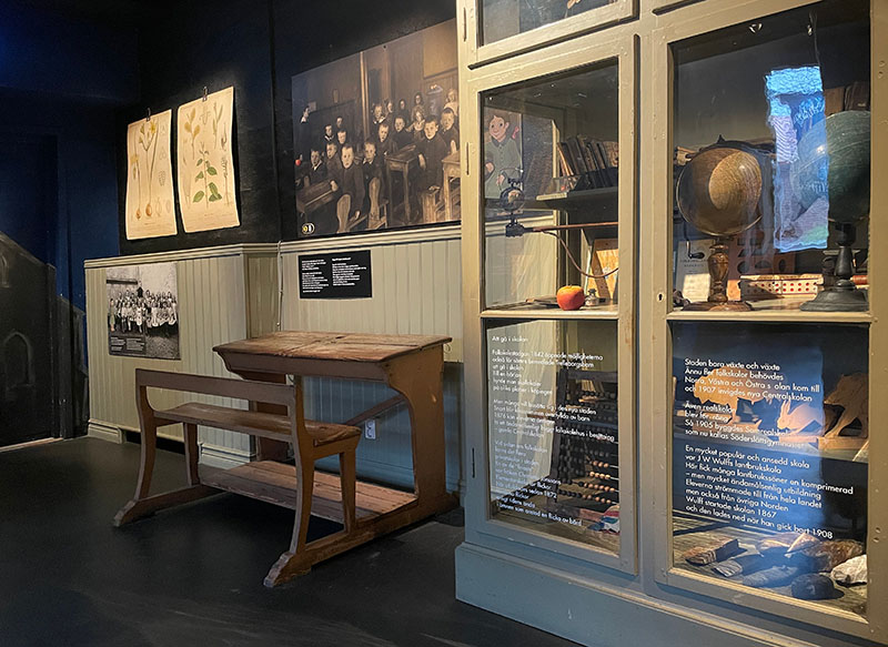 Vy från utställningen Stadslifv. En gammal skolbänk i trä finns intill ett glasskåp med äldre skolmaterial.