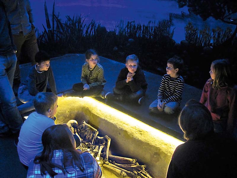 En vikingaklädd man och en vikingaklädd kvinna sitter vid ett bord utanför ett långhus.