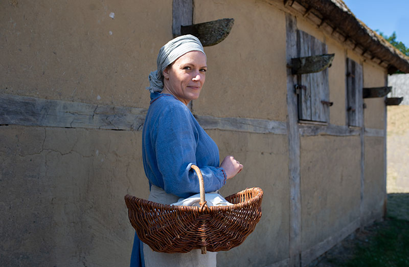 Medeltidsklädd kvinna står med en korg på armen framför et medeltida, lerklinat hus.