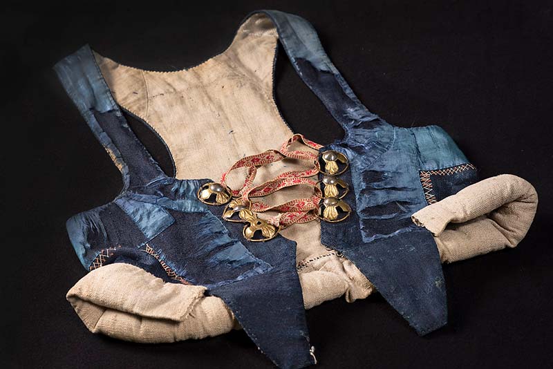 Ett klädesplagg som ser ut som en väst i blått, och fodrad med oblekt linnetyg. Med silverspännen och rött-vitt band.