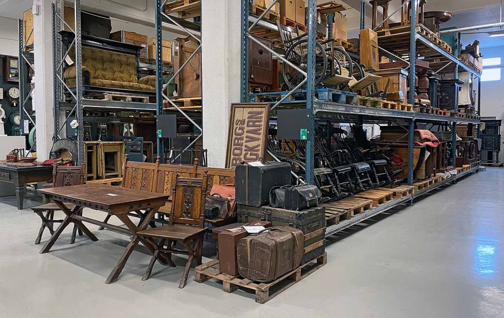 Hyllor med många föremål, i förgrunden bord, stolar och resväskor.