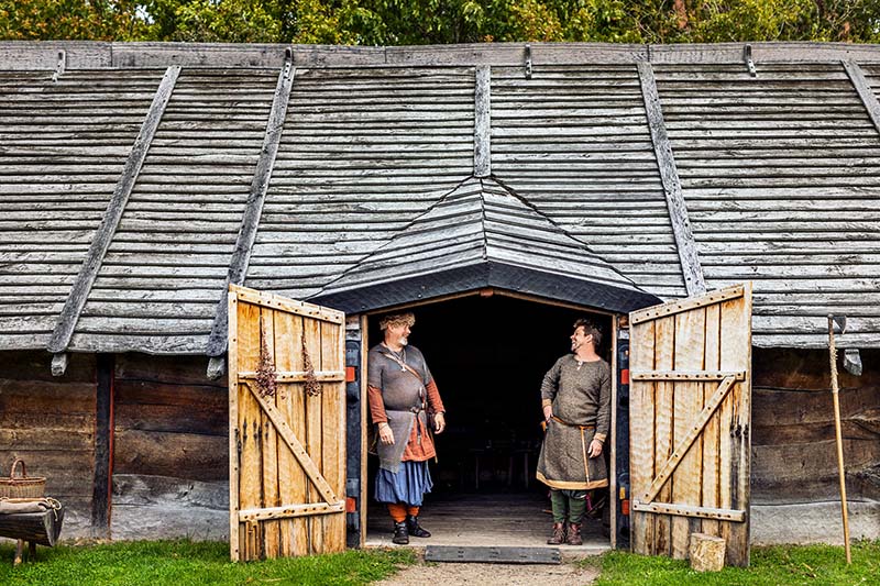 Två vikingaklädda män står i dörröppningen till gårdens långhus.