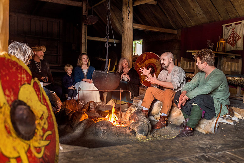 Flera besökare sitter runt en tänd eldstad inuti ett vikingatida långhus. De lyssnar på en vikingaklädd sagoberättare.