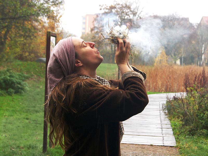 Siluett av kvinna i vikingakläder. Hon står ute i grönskan och blåser liv i glödande hö.
