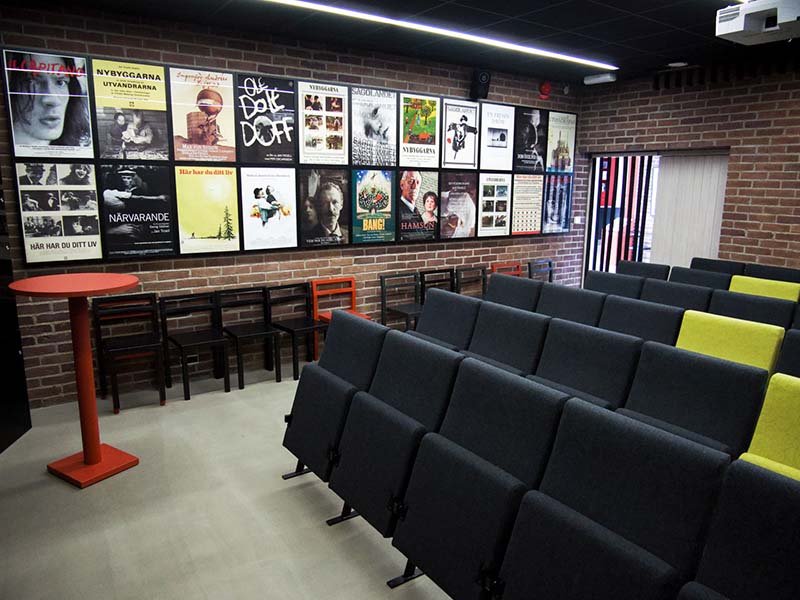 Room with rows of gray foldable chairs and black and red chairs next to a brown brick wall with film posters.