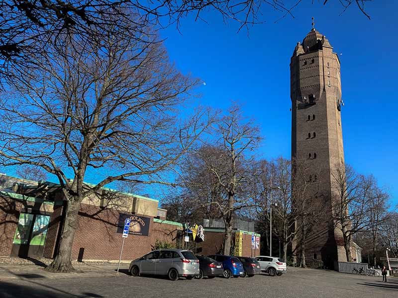 A brown brick building located next to a tall brick building near the parking area with occasional trees.