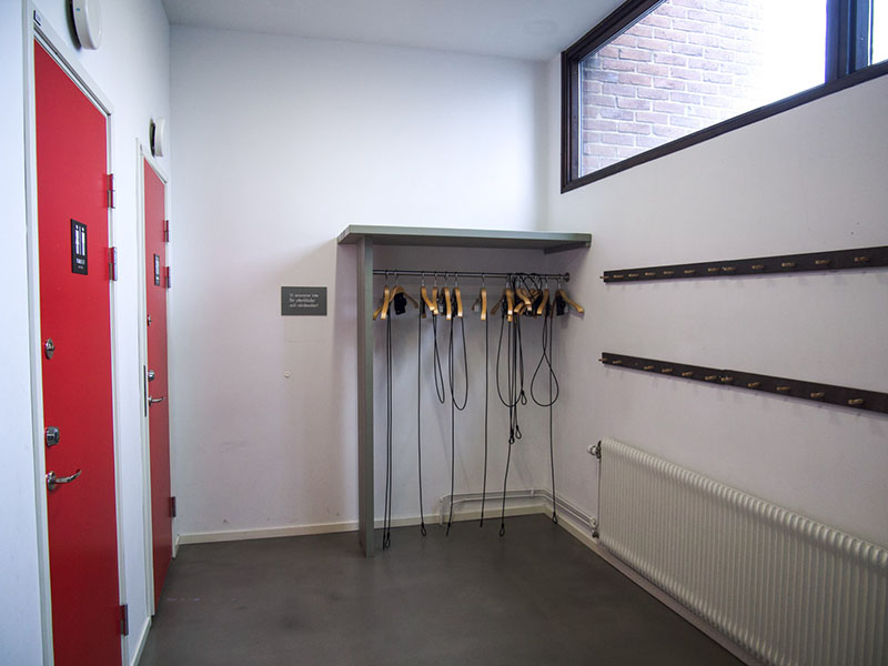 Bright room with white walls, gray floor, and two red doors with toilet symbols. Clothes hangers along the walls and windows at the ceiling level.