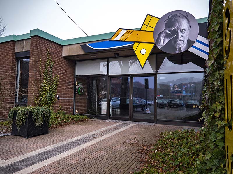 A tactile guiding strip leads to a brown brick building with glass doors. Vegetation and a sign on the facade depicting a person looking into a camera lens.