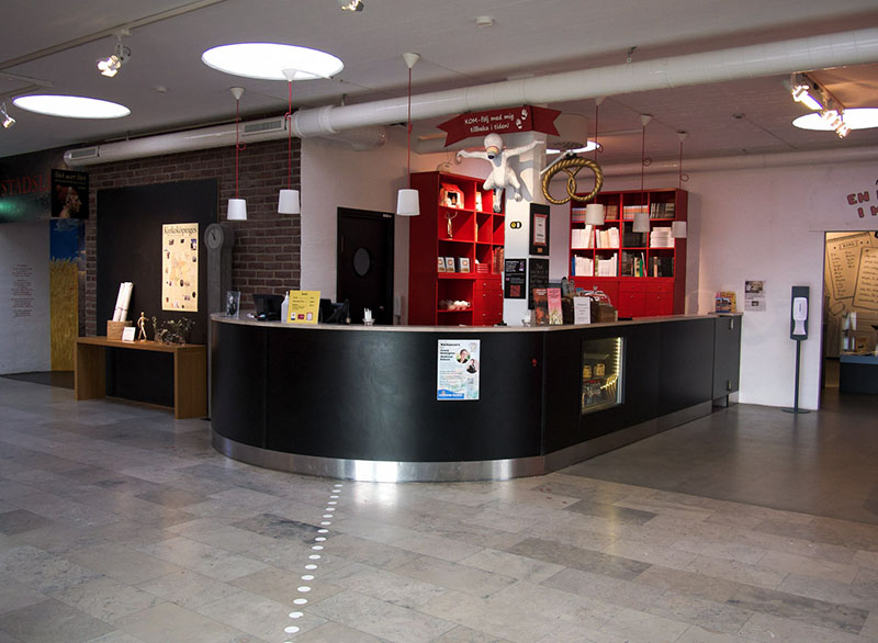 A black, high reception desk in an open room with a gray floor and white contrasting markings. A figure in the ceiling, red bookshelves, and various signs with information.