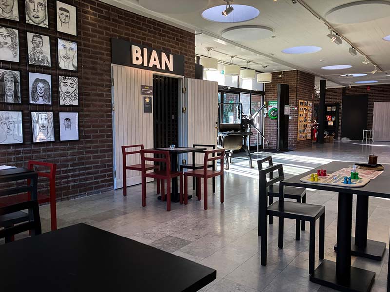 A bright open room with brick walls and a gray floor. Black and red tables and chairs with games. A doorway with a sign that reads "Bian." Artwork on the walls. An old-fashioned printing press, glass doors, and postcards in the background.
