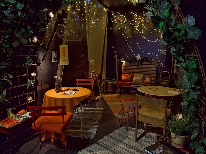 Dimly lit room with warm lights and string lights hanging from the ceiling. There are two yellow round tables with yellow and red chairs. Carpets cover the floor, and there is a bench against the wall with cushions and artificial plants. Outside the room, there is vegetation in an arched structure.