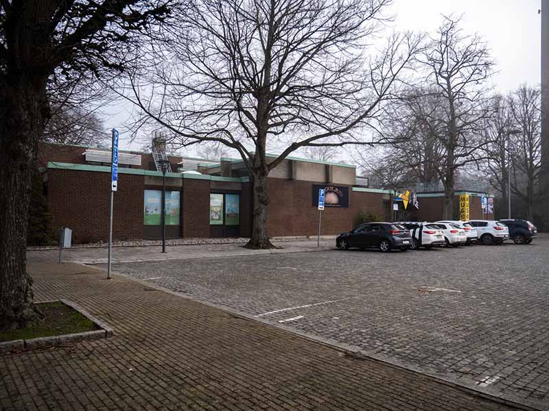 A designated accessible parking area with signage, adjacent to the general parking area with parked cars. In the background, there is a brown brick building and trees.