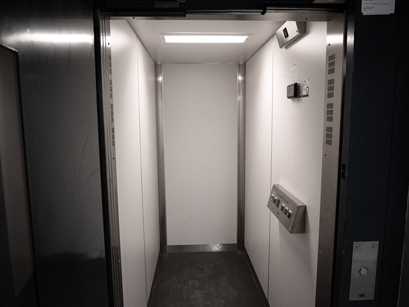 Inside of a narrow elevator, there are white walls and a gray floor.