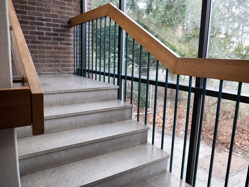 The gray staircase has a wooden handrail along the left side against a white wall. On the right side, there is a brick wall and a window facing a shrubbery straight ahead.