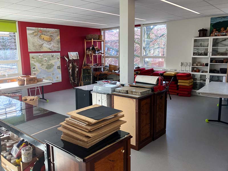 The large room has a light gray floor, white and red walls. There are benches, shelves, and tables along the walls and near a white slim pillar in the middle of the room. Paint, various tools, cushions, and materials are located near the benches and shelves.