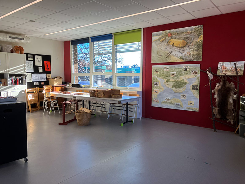 A large room with a light gray floor, white and red walls, and large windows. It contains tables with chairs and wooden tools on the floor and tables.