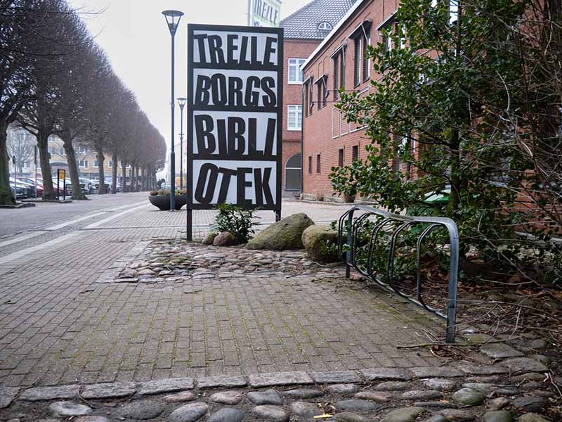 Bicycle rack located on a paved area next to a shrubbery. There is a large white sign with black text that reads “Trelleborgs Bibliotek” (Trelleborg Library). In the background, you can see a row of trees and red brick buildings.