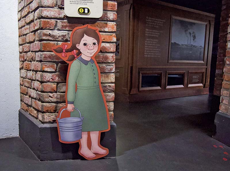 A cartoon girl wearing a green dress, brown hair with two pigtails, holding a metal mucket. It is printed on a disc mounted on a brIck wall.