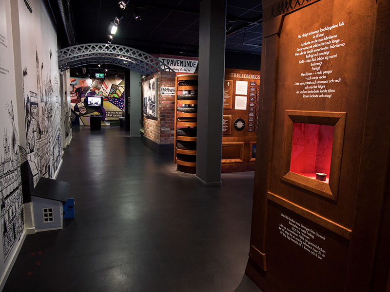 A large room with walls of various colors and a black floor. There is a small blue house on the floor next to the left wall. The display cases are made of wood with white text and objects.