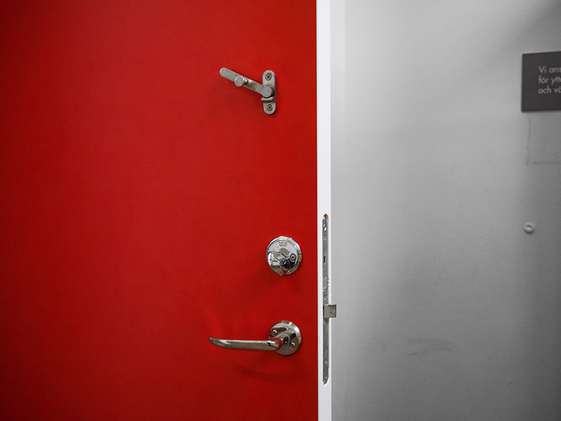 Close-up of a red door against a white wall. Handle and deadlock, as well as a hasp.