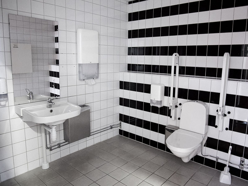 Large bathroom with gray floor and black-and-white walls. Accessible toilet, toilet paper, and toilet brush to the right. Sink with faucet and mirror, paper towels for drying, and a trash can to the left.