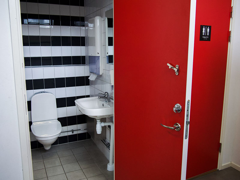 Open, red door revealing a bathroom with a toilet, sink, and paper towels. Gray floor and black-and-white walls.