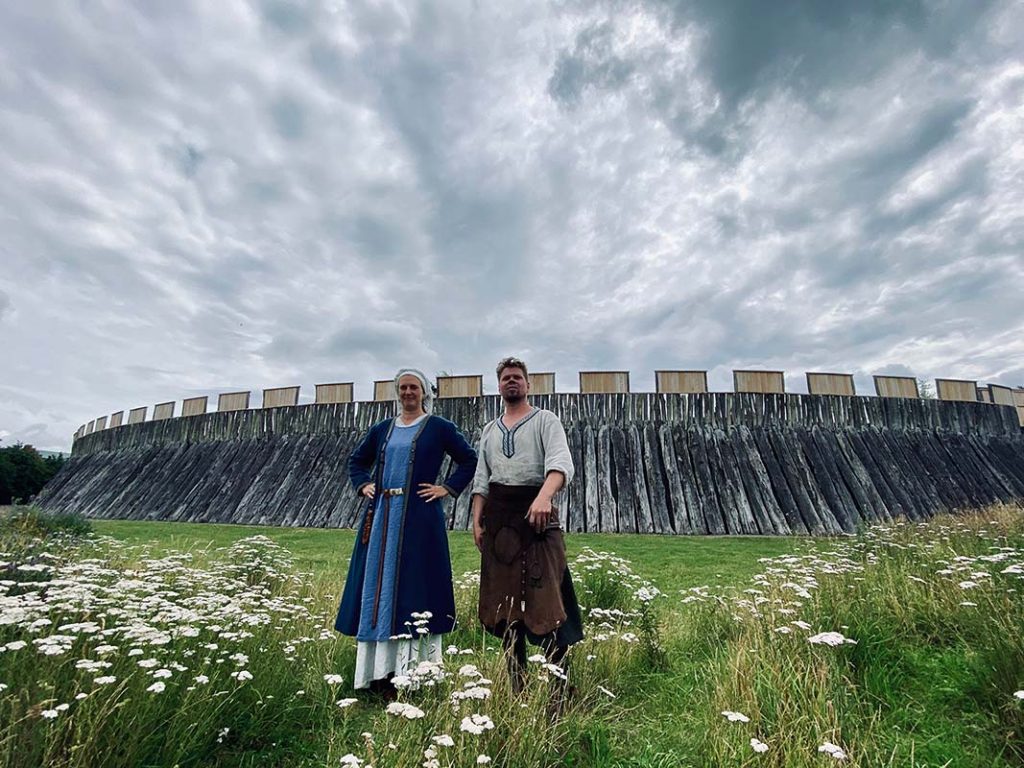 Två vikingaklädda personer framför Trelleborgen, som är cirkelrund borg byggd av ekstockar.