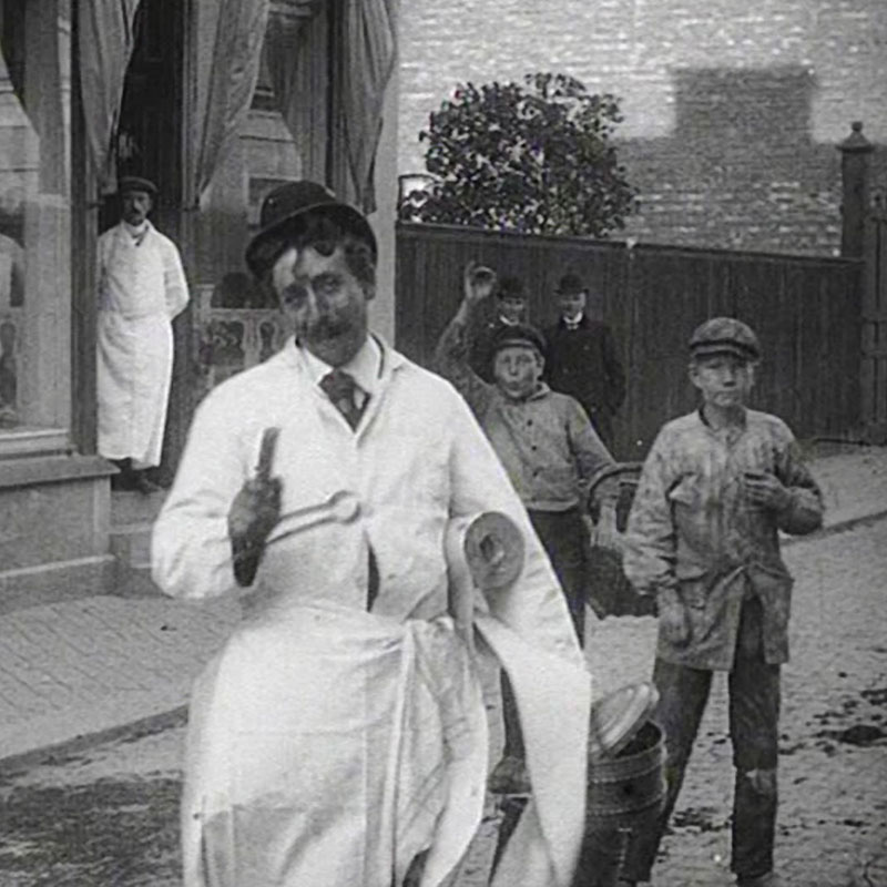 Filmruta från äldre svartvit film. En korvförsäljare står på en gata. Ett par pojkar står bakom honom. Tre män tittar på.