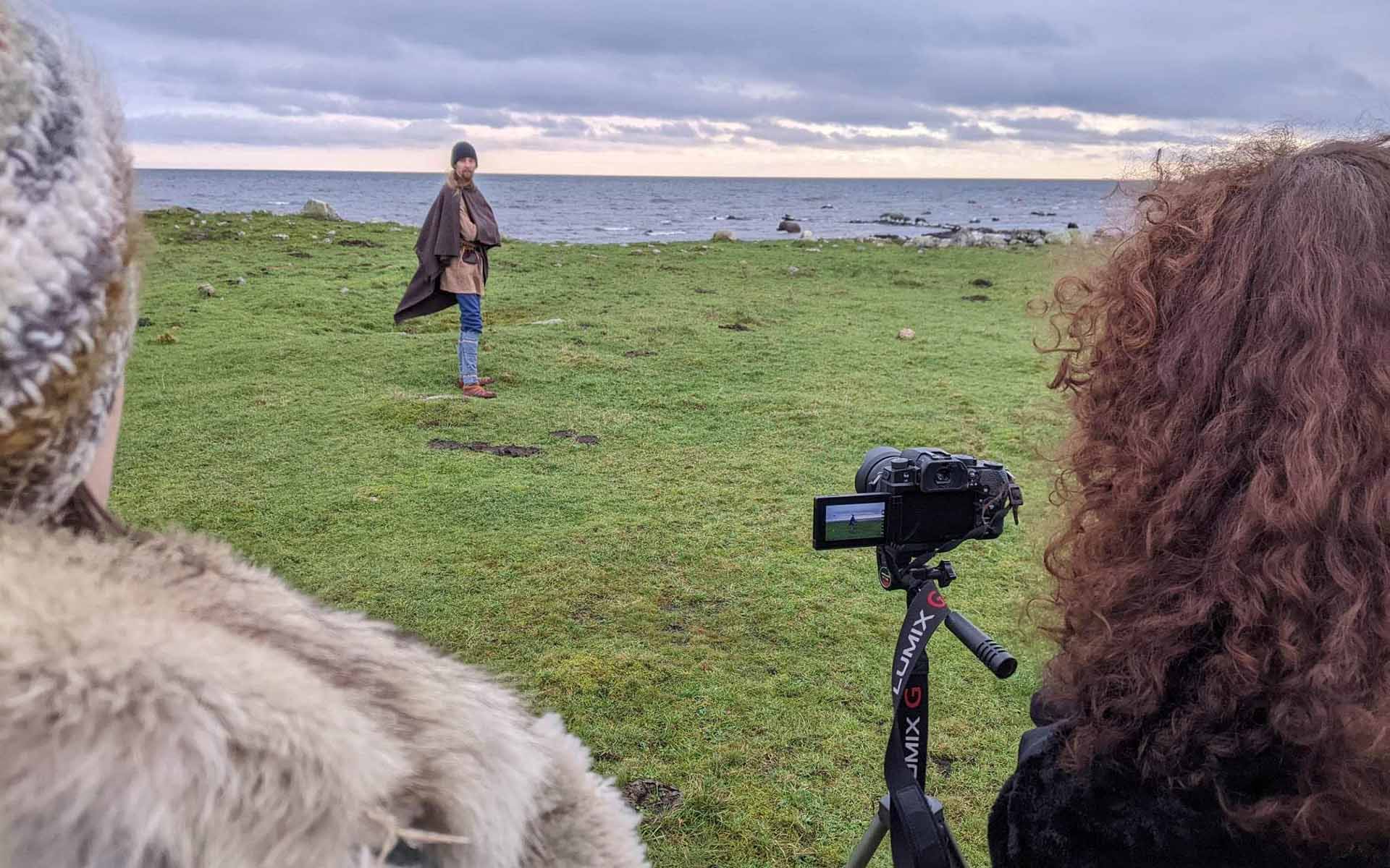 Två personer filmar en man i vikingakläder. De tvår på en äng där man ser havet i bakgrunden.