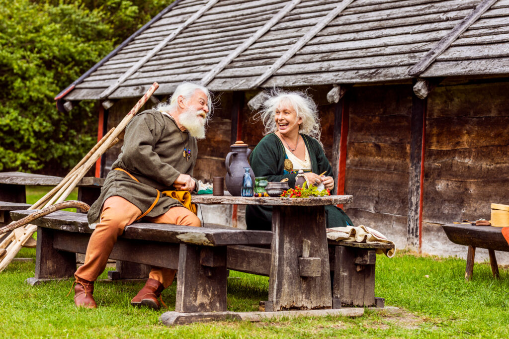 Skrattande äldre par i vikingatida kläder sitter utanför långhuset på Trelleborgen.