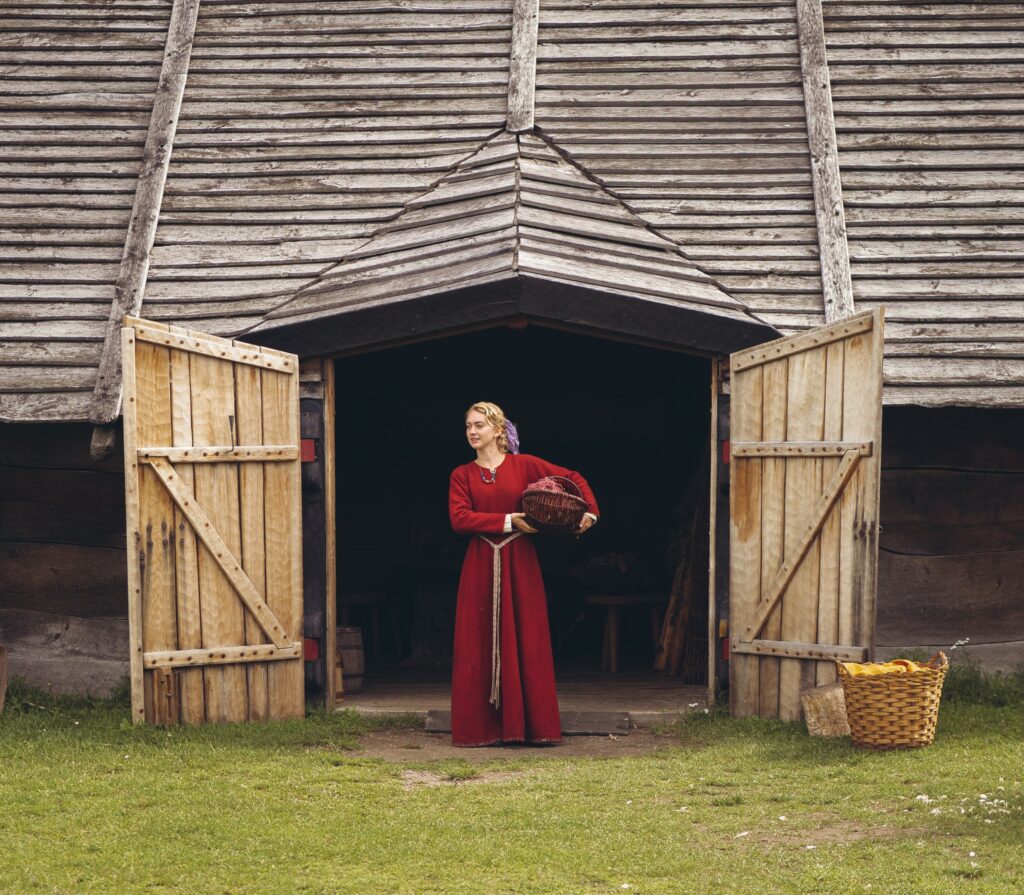 Kvinna i röd klänning står i dörröppningen till ett vikingatida långhus, på Trelleborgen