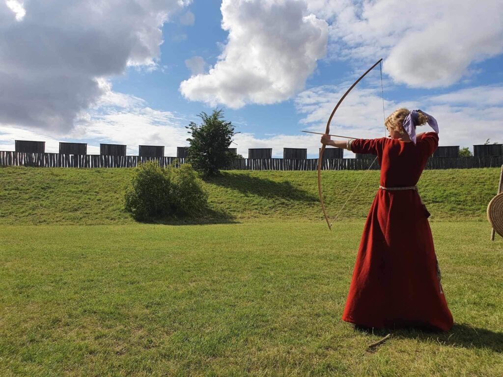 Kvinna i vikingatida klänning skjuter pilbåge. I bakgrunden ser man Trelleborgen.