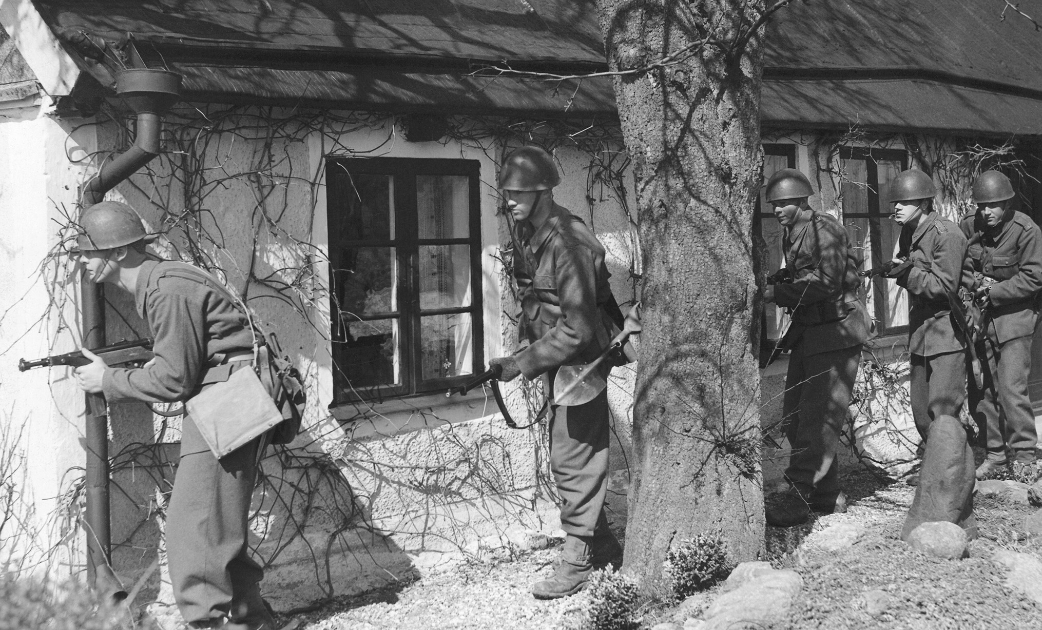 Soldater i uniform tränar , april 1942. De gömmer sig bakom ett ett hus, beväpnade med gevär.