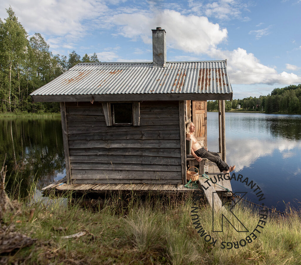 Liten bastu av trä som ligger vid en sjö. En man sitter med slutna ögon bredvid bastun. Foto Aija Svensson