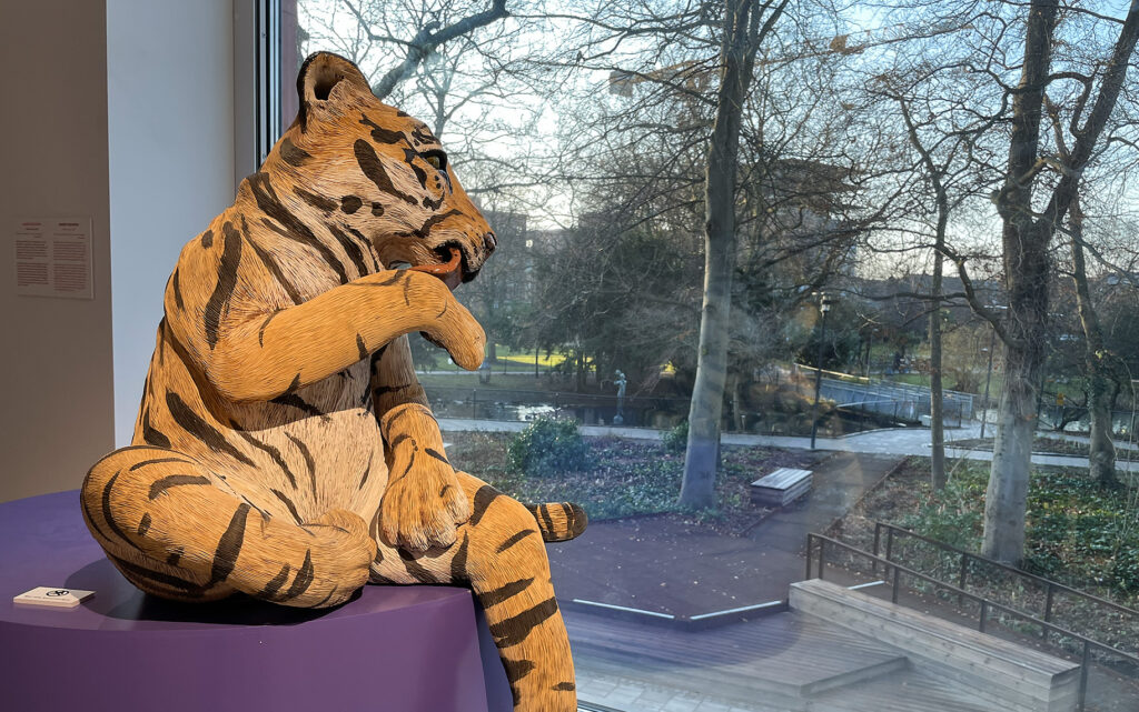 Skulptur av en tiger som sitter på ett lila podium. Bredvifd finns ett fönster med utsikt över Stadsparken. Skulptur av Margit Brundin.