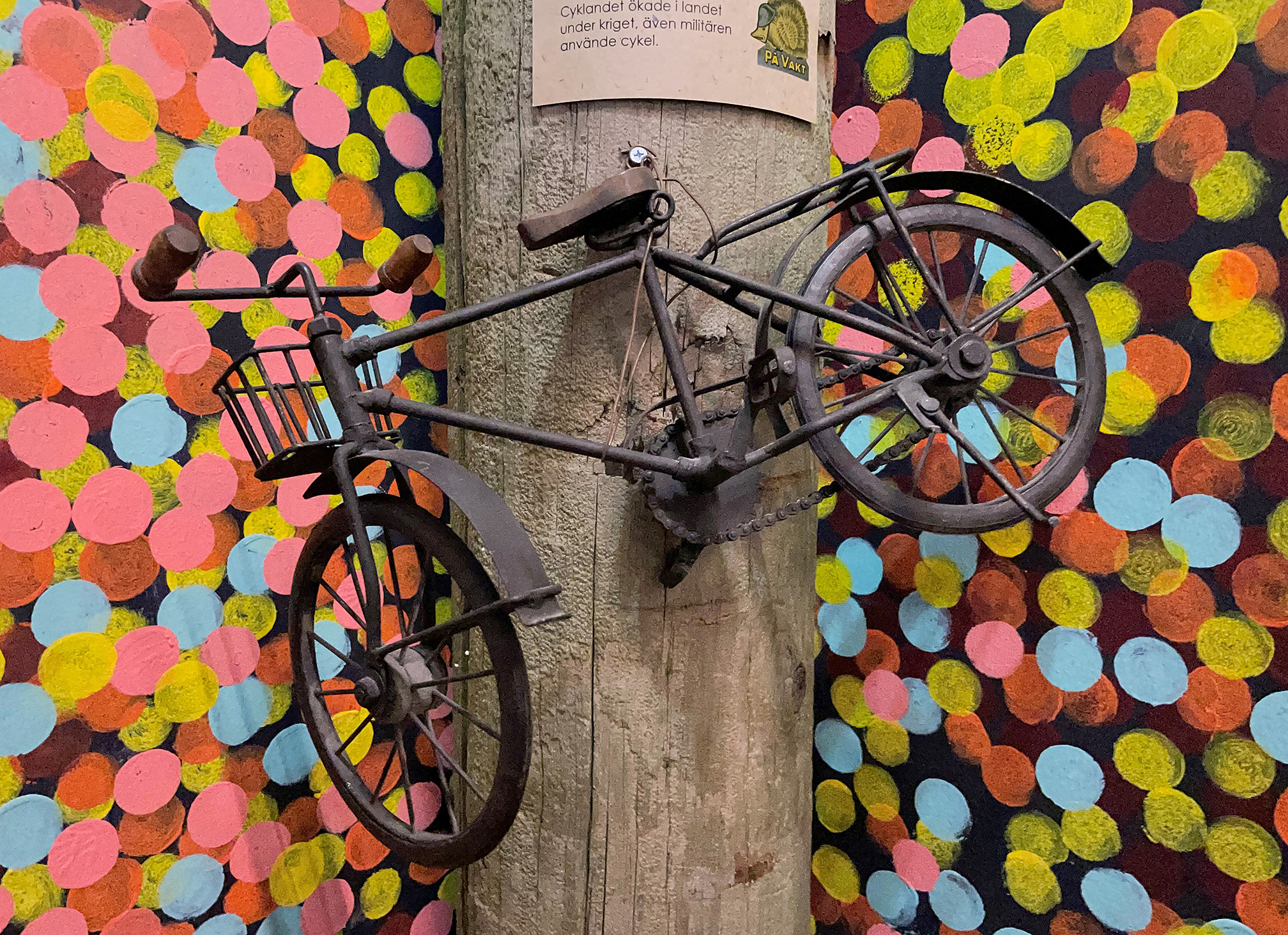 Liten cykel sitter fäst på en stolpe. Prickar i många färger målade på väggen bakom.