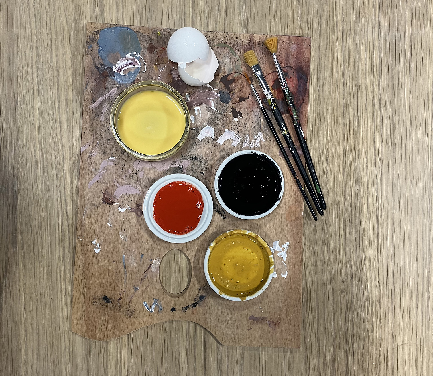 penslar och burkar med temperafärg i rött, svart och två gula nyanser står på en palett.