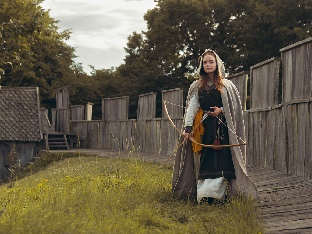 Vikingaklädd kvinna med pilbåge. Trelleborgen i bakgrunden.